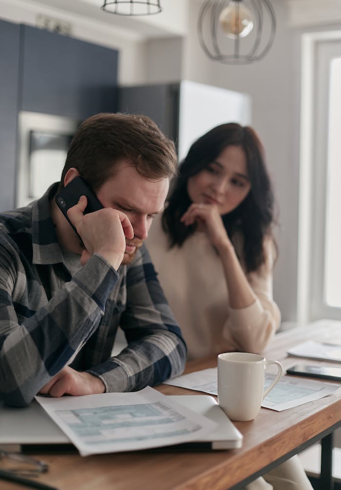 A worried couple sits at a table with bills and a phone, planning their finances.